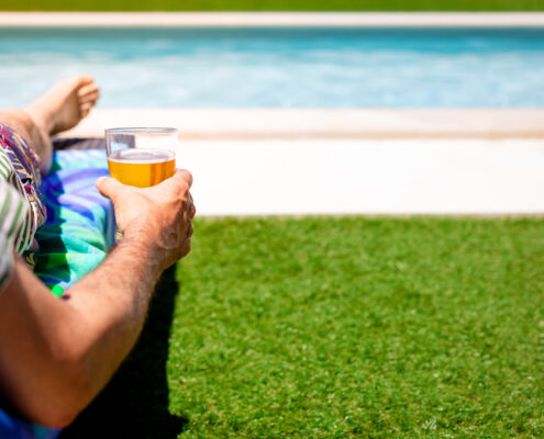 Mann, der sich mit einem Bier in der Hand, auf einer Sonnenliege im Garten am Pool entspannt.