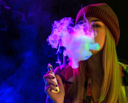 Eine junge Frau trägt eine rote Strickmütze und dampft eine E-Zigarette, umgeben von buntem lila und pinkem Dampf vor einem dunklen Hintergrund.