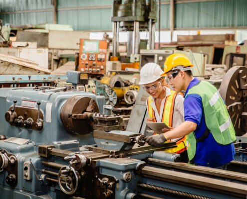 Zwei Techniker in Schutzkleidung überprüfen eine industrielle Maschine in einer Werkstatt und besprechen die Anwendung von Druckluftschmiergeräten zur optimalen Pflege.