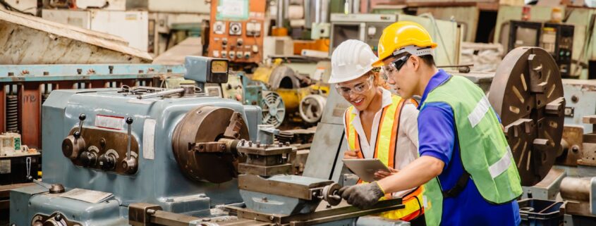 Zwei Techniker in Schutzkleidung überprüfen eine industrielle Maschine in einer Werkstatt und besprechen die Anwendung von Druckluftschmiergeräten zur optimalen Pflege.