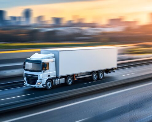Ein weißer Lkw fährt bei Sonnenaufgang auf einer mehrspurigen Autobahn mit verschwommenem Stadthintergrund – symbolisiert moderne Logistik und Bewegung.
